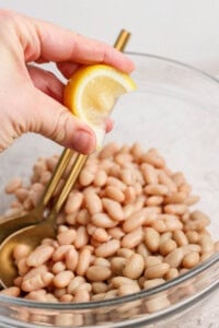 A hand squeezing a lemon wedge over a bowl of white beans, with a gold spoon resting inside the bowl.
