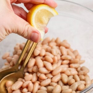 A hand squeezing a lemon wedge over a bowl of white beans, with a gold spoon resting inside the bowl.