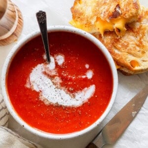 A bowl of tomato soup swirled with cream and pepper sits beside a grilled cheese sandwich, accompanied by a knife and a striped napkin.