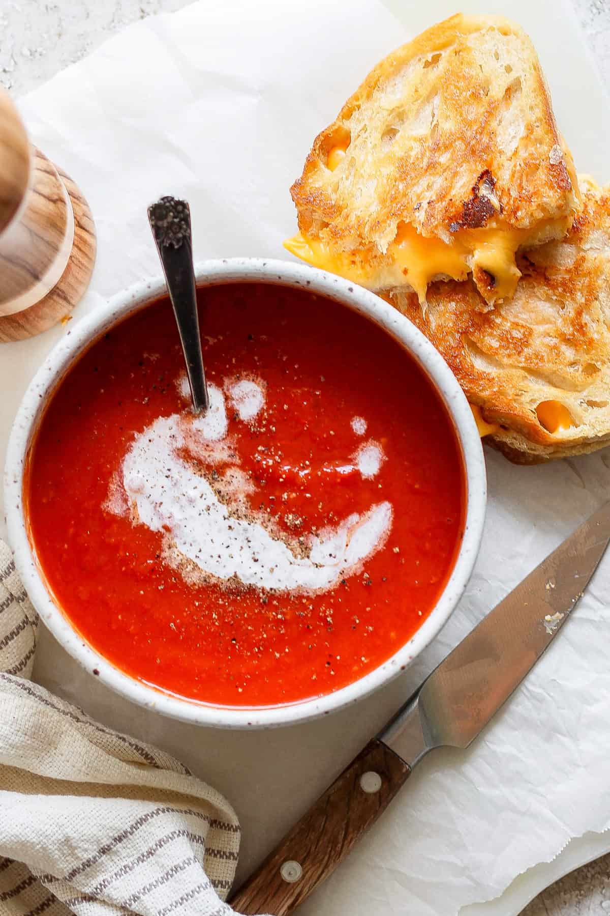 A bowl of tomato soup swirled with cream and pepper sits beside a grilled cheese sandwich, accompanied by a knife and a striped napkin.