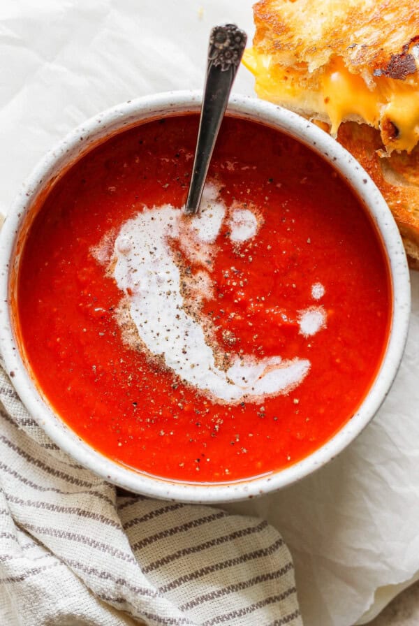 A bowl of creamy tomato soup topped with pepper, paired perfectly with a melty grilled cheese sandwich. A spoon rests invitingly in the bowl, while a striped cloth napkin sits nearby.