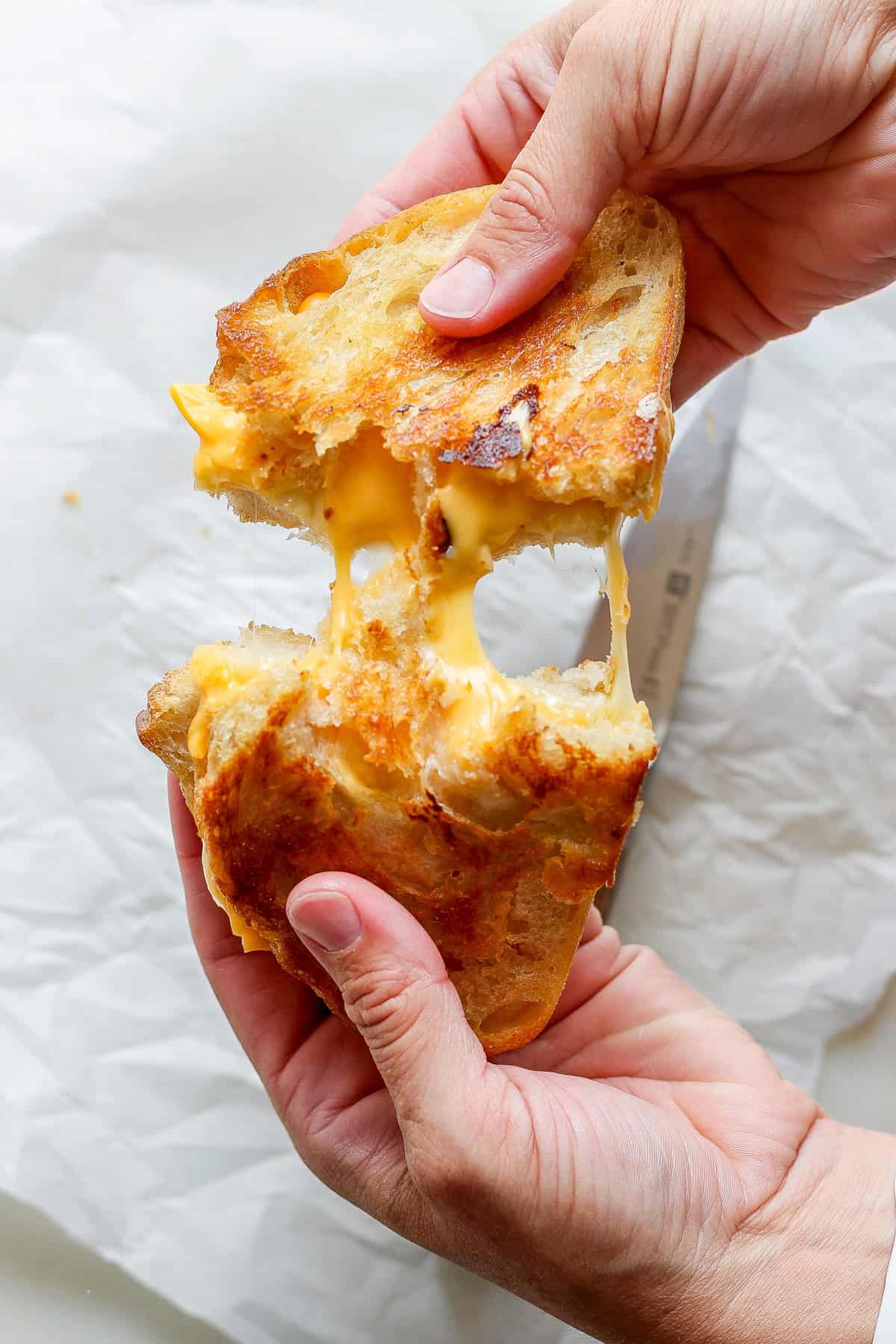 Hands pulling apart a grilled cheese sandwich, with melted cheese stretching between the toasted bread slices, perfectly paired with a warm bowl of tomato soup.