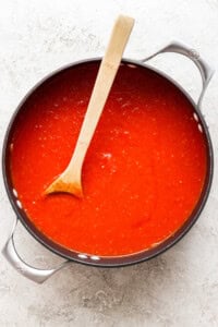 A pot filled with rich, red tomato sauce and a wooden spoon resting inside evokes the comforting aroma of grilled cheese and tomato soup on a light-colored surface.