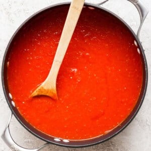 A pot filled with rich, red tomato sauce and a wooden spoon resting inside evokes the comforting aroma of grilled cheese and tomato soup on a light-colored surface.