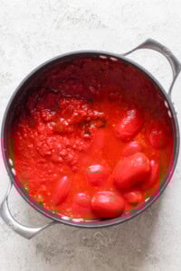 A black pot filled with whole and crushed tomatoes in tomato sauce, reminiscent of a comforting bowl of tomato soup, sits on a light gray surface. Perfect for pairing with a crispy grilled cheese sandwich.