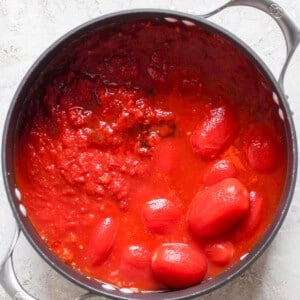 A black pot filled with whole and crushed tomatoes in tomato sauce, reminiscent of a comforting bowl of tomato soup, sits on a light gray surface. Perfect for pairing with a crispy grilled cheese sandwich.