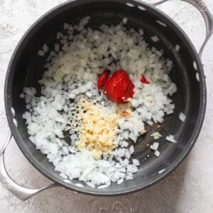 Chopped onions, minced garlic, and dollops of tomato paste in a black pot on a light textured surface evoke the comforting aroma of grilled cheese and tomato soup.