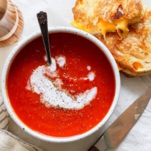 A bowl of rich tomato soup swirled with cream and pepper sits beside a perfect pairing: a crispy grilled cheese sandwich. A knife rests nearby on the table, alongside a neatly folded napkin, completing the classic duo's delicious presentation.