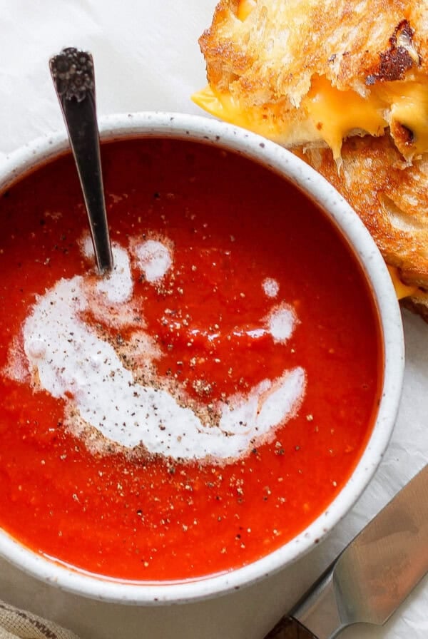 A bowl of rich tomato soup swirled with cream and pepper sits beside a perfect pairing: a crispy grilled cheese sandwich. A knife rests nearby on the table, alongside a neatly folded napkin, completing the classic duo's delicious presentation.