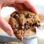 A hand holding a paleo chocolate chip cookie with a bite taken out, next to a glass of milk.