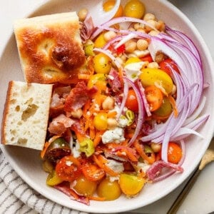 A salad with cherry tomatoes, red onion, carrot shreds, bacon, cheese, and chickpeas, accompanied by two bread pieces. Dressing is in a small cup nearby.