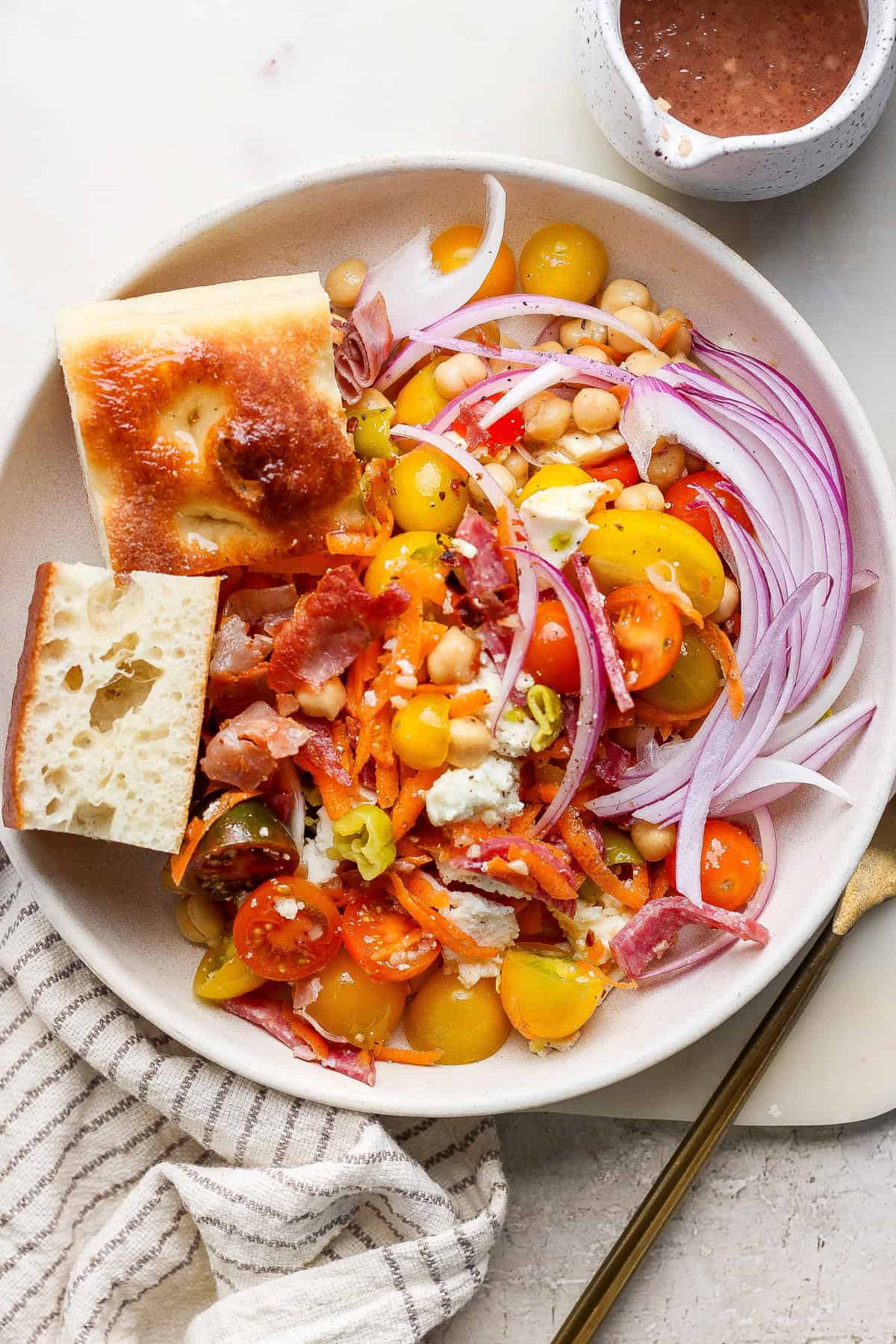 A salad with cherry tomatoes, red onion, carrot shreds, bacon, cheese, and chickpeas, accompanied by two bread pieces. Dressing is in a small cup nearby.