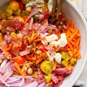 A salad bowl with shredded carrots, sliced red onions, cherry tomatoes, chickpeas, feta cheese, chopped bacon, and salami. Two forks are in the bowl.