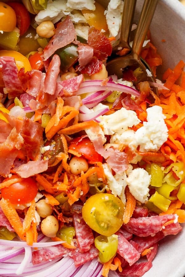 A salad bowl with shredded carrots, sliced red onions, cherry tomatoes, chickpeas, feta cheese, chopped bacon, and salami. Two forks are in the bowl.