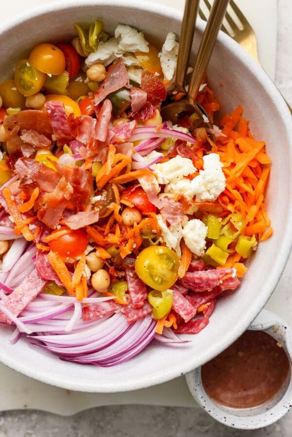 A bowl of salad with sliced red onions, cherry tomatoes, chickpeas, shredded carrots, crumbled cheese, and cured meats. Two forks are placed in the bowl. A small bowl of dressing is beside it.