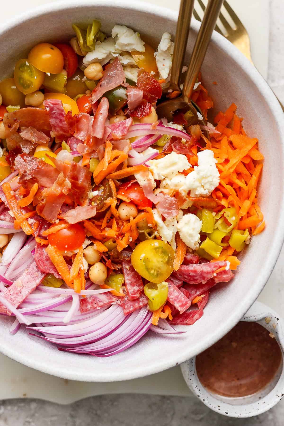 A bowl of salad with sliced red onions, cherry tomatoes, chickpeas, shredded carrots, crumbled cheese, and cured meats. Two forks are placed in the bowl. A small bowl of dressing is beside it.