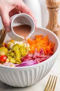 A hand pours dressing onto a salad with tomatoes, chickpeas, salami, red onions, pepperoncini, shredded carrots, and feta in a bowl. A fork and pepper grinder are nearby.