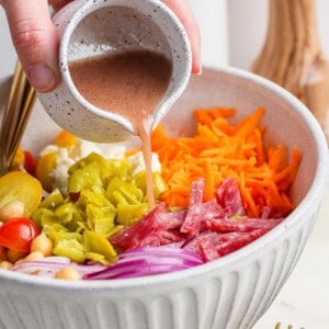 A hand pours dressing onto a salad with tomatoes, chickpeas, salami, red onions, pepperoncini, shredded carrots, and feta in a bowl. A fork and pepper grinder are nearby.