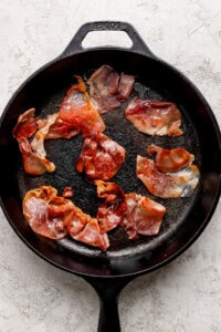 Slices of cooked prosciutto in a black cast iron skillet on a light textured surface.