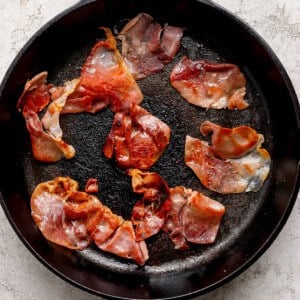 Slices of cooked prosciutto in a black cast iron skillet on a light textured surface.