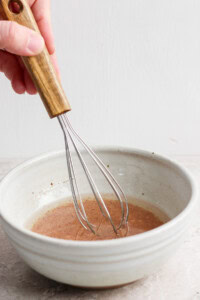 A hand using a wooden-handled whisk to stir liquid in a ceramic bowl.