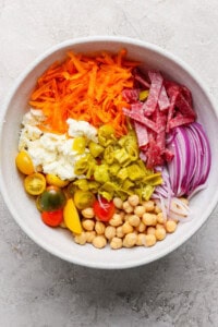 Bowl with shredded carrots, sliced salami, red onions, chickpeas, cherry tomatoes, mozzarella, and chopped green peppers.
