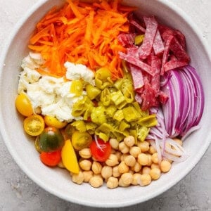 Bowl with shredded carrots, sliced salami, red onions, chickpeas, cherry tomatoes, mozzarella, and chopped green peppers.