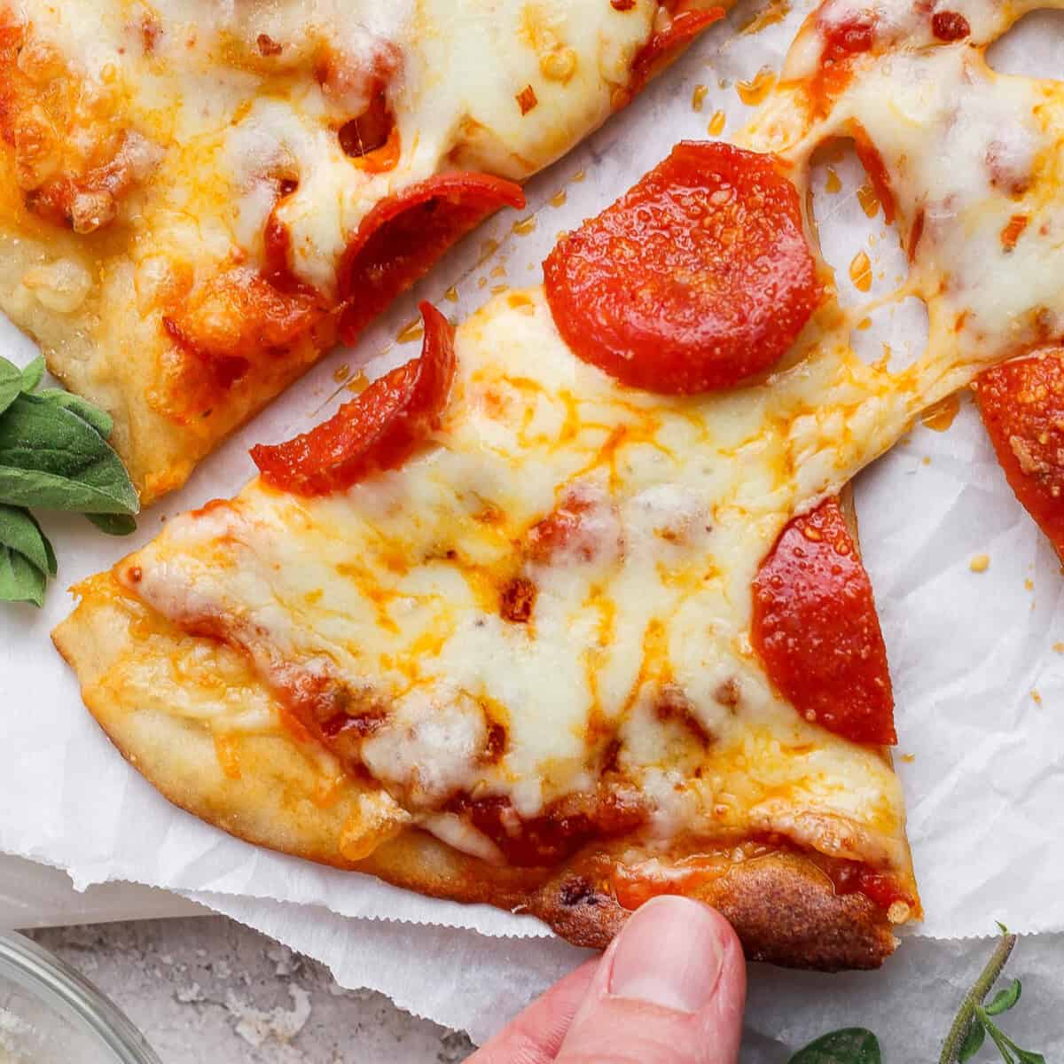 A hand holding a slice of pepperoni pizza with melted cheese on a white parchment background.