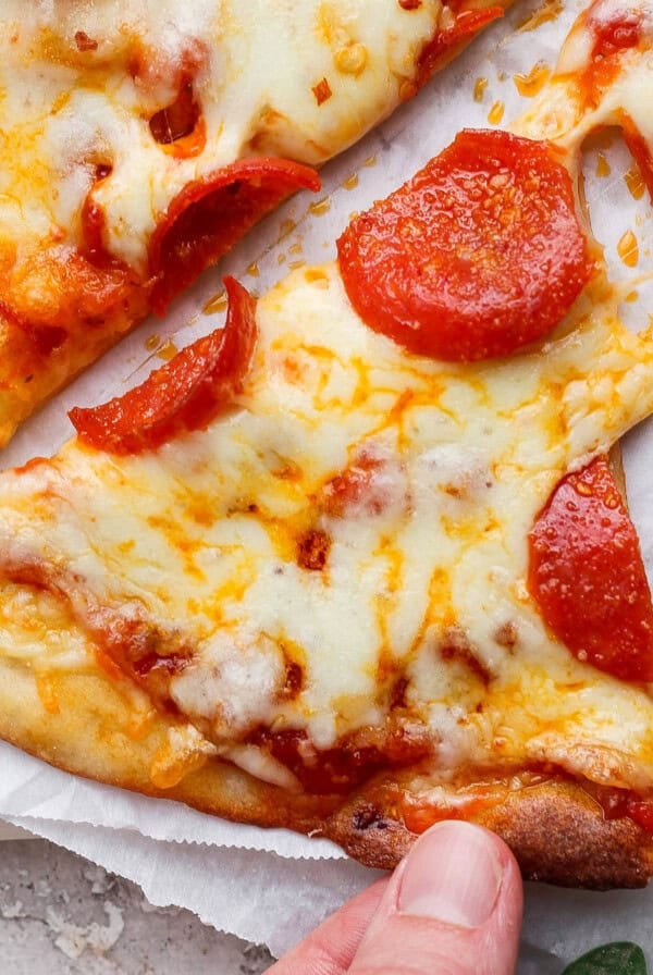 A hand holding a slice of pepperoni pizza with melted cheese on a white parchment background.