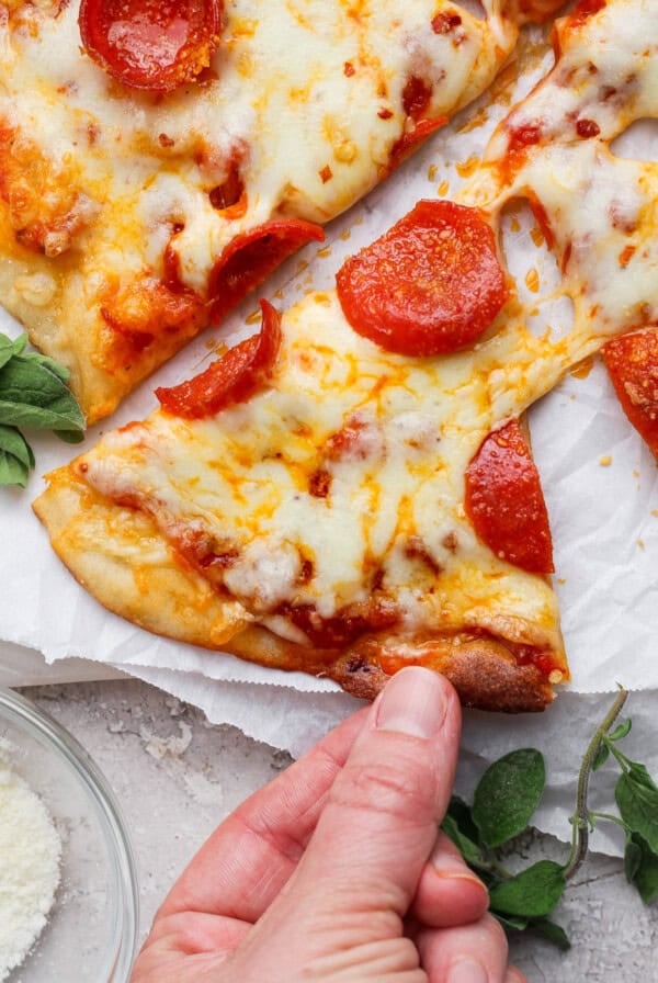 A hand holding a slice of pepperoni pizza topped with melted cheese, with oregano and a small bowl of grated cheese nearby.