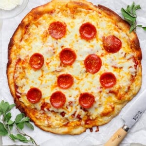 A pepperoni pizza on parchment paper with a knife beside it, garnished with herbs. A small bowl of grated cheese is in the top-left corner.