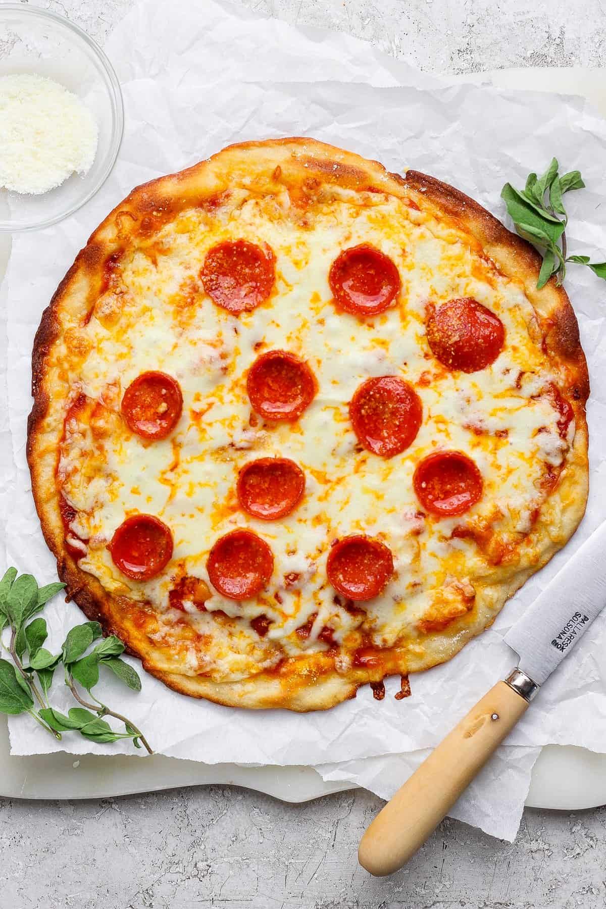 A pepperoni pizza on parchment paper with a knife beside it, garnished with herbs. A small bowl of grated cheese is in the top-left corner.