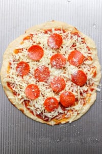 Unbaked pizza with pepperoni slices, grated cheese, and sauce on a round dough placed on a textured metal surface.