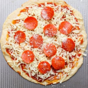 Unbaked pizza with pepperoni slices, grated cheese, and sauce on a round dough placed on a textured metal surface.