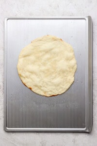 A round, uncooked pizza dough is placed on a metal baking sheet with a textured surface.