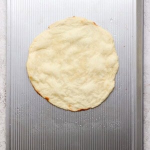 A round, uncooked pizza dough is placed on a metal baking sheet with a textured surface.