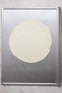 A round, uncooked pizza dough is placed on a textured metal baking sheet.
