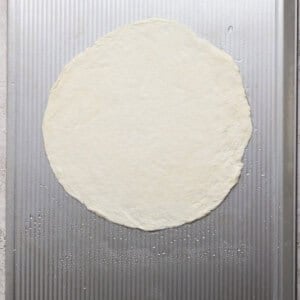 A round, uncooked pizza dough is placed on a textured metal baking sheet.