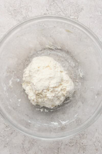A ball of dough in a clear glass bowl on a light textured surface.