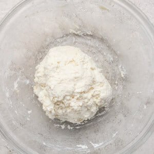 A ball of dough in a clear glass bowl on a light textured surface.