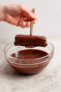 A hand holds chopsticks dipping a chocolate-covered rectangular treat into a glass bowl of melted chocolate.