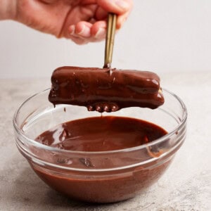 A hand holds chopsticks dipping a chocolate-covered rectangular treat into a glass bowl of melted chocolate.