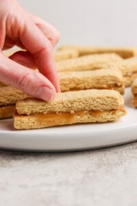 A hand picks up a peanut butter sandwich cookie from a white plate. More cookies are visible in the background.