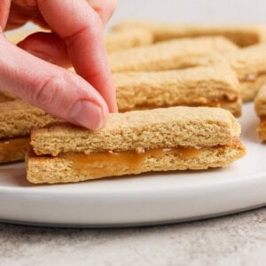 A hand picks up a peanut butter sandwich cookie from a white plate. More cookies are visible in the background.