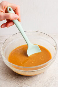A hand stirs brown gravy in a clear glass bowl with a light green spatula on a light surface.