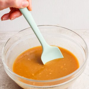 A hand stirs brown gravy in a clear glass bowl with a light green spatula on a light surface.