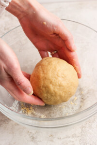Hands shaping a ball of dough in a clear glass bowl on a light-colored surface.