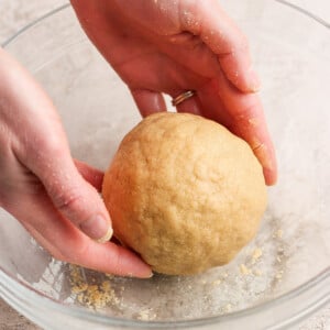 Hands shaping a ball of dough in a clear glass bowl on a light-colored surface.