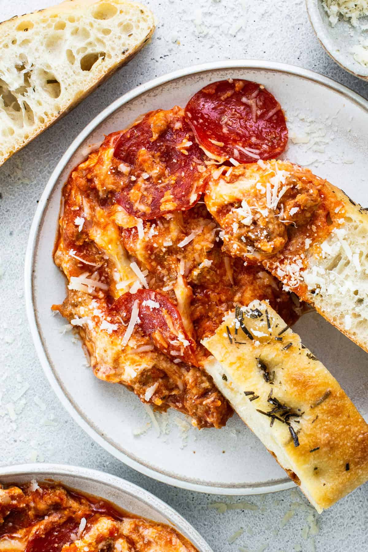 A plate of pepperoni pizza dip topped with cheese, accompanied by slices of crusty bread and rosemary focaccia.