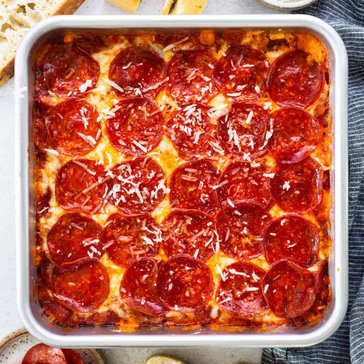 Square pan of pepperoni pizza with a golden crust, melted cheese, and sliced pepperoni, topped with grated cheese, surrounded by bread slices and a striped napkin.